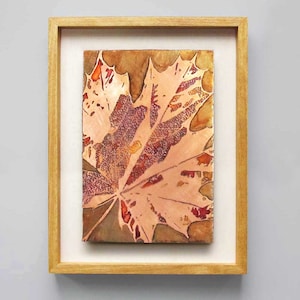 May include: A framed artwork featuring a close-up of a single maple leaf with intricate details. The leaf is a vibrant mix of orange, red, and brown hues, creating a beautiful autumnal scene.