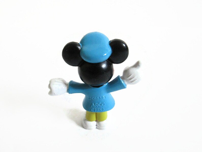 May include: A small Mickey Mouse figurine in a light blue outfit with a matching hat. The figure has black ears and a black face, with white gloves and yellow pants. The figurine is marked "Disney" and "ARGO CHINA".
