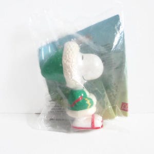 May include: A Snoopy figurine in a clear plastic bag. Snoopy is wearing a green hat and jacket with white trim and red accents. The figurine is white with red shoes. A blue and green background is visible.