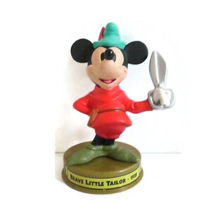 May include: A Mickey Mouse figurine dressed as the Brave Little Tailor, wearing a red tunic, green hat, and green shoes. The character holds a silver-colored pair of scissors in one hand. The base is circular and inscribed with Brave Little Tailor 1938.