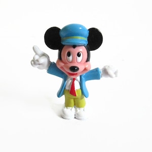 May include: A small, colorful Mickey Mouse figurine. Mickey is wearing a blue conductor's hat and jacket, a red tie, yellow pants, and white gloves and shoes. He is pointing with his right hand.