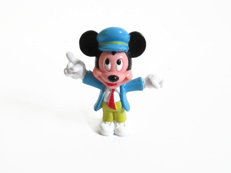 May include: A small, colorful Mickey Mouse figurine. Mickey is wearing a blue conductor's hat and jacket, a red tie, yellow pants, and white gloves and shoes. He is pointing with his right hand.