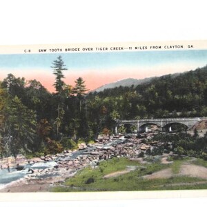 May include: A vintage postcard featuring a colorized landscape of Saw Tooth Bridge over Tiger Creek, 11 miles from Clayton, GA. The scene depicts a stone arch bridge spanning a rocky creek surrounded by dense green forest and a soft sunset sky.