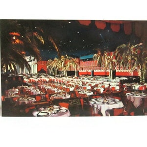May include: A vintage postcard depicting a large dining hall with rows of round tables covered in white tablecloths, surrounded by red chairs and palm trees. The room features a dark ceiling with recessed lighting and red wall accents.