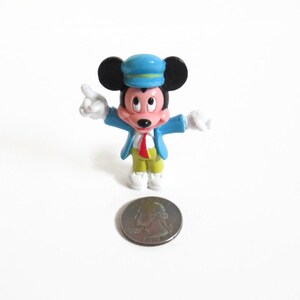 May include: A small Mickey Mouse figurine wearing a blue conductor's hat and jacket, red tie, yellow pants, and white shoes. The figurine is standing next to a quarter coin on a white surface.