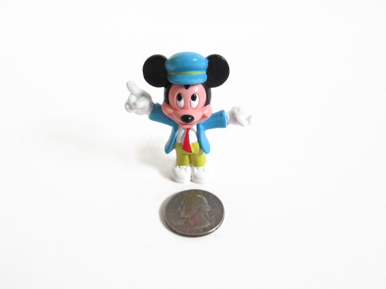 May include: A small Mickey Mouse figurine wearing a blue conductor's hat and jacket, red tie, yellow pants, and white shoes. The figurine is standing next to a quarter coin on a white surface.