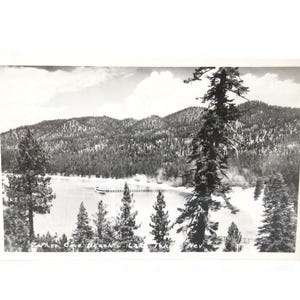 May include: Black and white vintage postcard featuring a scenic view of a lake surrounded by mountains and evergreen trees. A pier extends into the lake, and the text "Zephyr Cove Beach, Lake Tahoe, Nev." is visible.