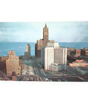 May include: Vintage postcard featuring a cityscape with tall buildings and a body of water in the background. The buildings are of varying heights and architectural styles, with a clear sky above. The postcard has a slightly faded appearance.