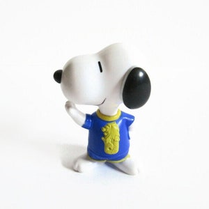 May include: A white Snoopy figurine with black ears and a black nose, wearing a blue shirt with a yellow emblem. The figurine is in a kneeling pose, with one arm raised.