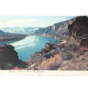 May include: Vintage postcard featuring a scenic view of the Mighty Columbia River in Washington. The image shows a wide river flowing between rocky cliffs and mountains under a blue sky. The postcard has a scalloped edge.