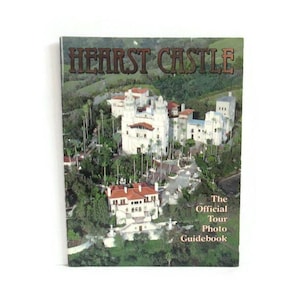 May include: A Hearst Castle tour guidebook featuring a photograph of the historic castle with white walls and red-tiled roofs, surrounded by lush green trees. The title "HEARST CASTLE" is displayed at the top, with "The Official Tour Photo Guidebook" below.