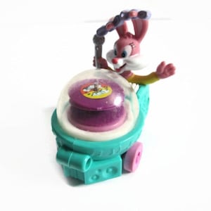 May include: A vintage toy featuring a pink bunny character with a white face and yellow gloves. The bunny is holding a purple and clear dome-shaped object with a teal base and pink wheels. The toy has a retro aesthetic.