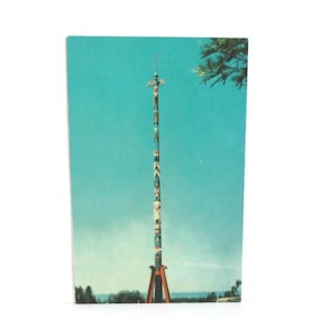 May include: Vintage postcard featuring a tall, colorful totem pole against a clear, turquoise sky. The totem pole is intricately carved with various figures and symbols. The base of the pole rests on a red wooden structure, with a few trees and the horizon visible in the background.