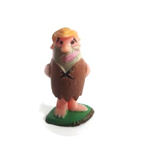 May include: A small plastic figurine of a cartoon character with blonde hair, a light skin tone, and a brown tunic featuring a black X-shaped detail on the chest. The character stands on a small, flat, green base with visible toes and feet.