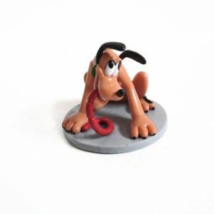 May include: A Pluto figurine, featuring the Disney cartoon dog in a playful pose. The figurine is primarily tan with black ears, a red tongue, and a green collar. It sits on a gray circular base.