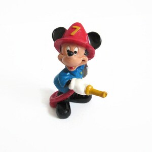 May include: A small figurine of Mickey Mouse dressed as a firefighter, wearing a red helmet with the number 7 in yellow, a blue jacket, and black shoes. The character is holding a white and yellow fire hose nozzle while standing in a pose.