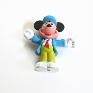 May include: A small, colorful figurine of a cartoon mouse character. The mouse is wearing a blue jacket, a red tie, yellow pants, and a blue hat. The mouse has black ears, a pink face, and white gloves and shoes. The figurine is standing with its arms outstretched.