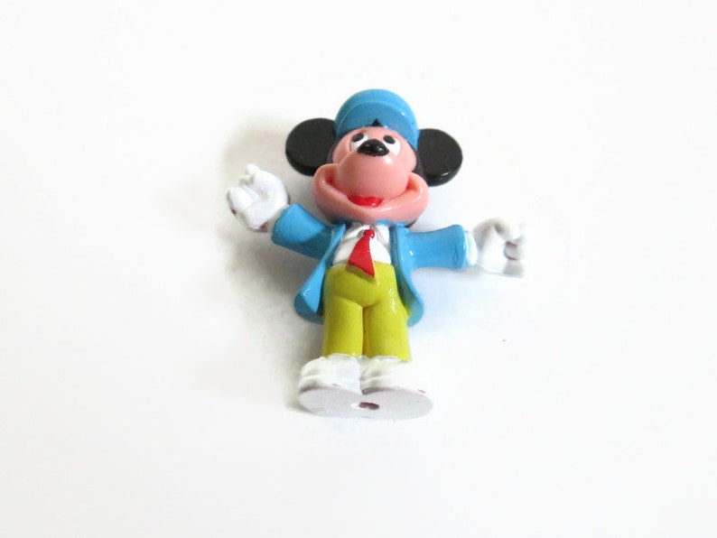 May include: A small, colorful figurine of a cartoon mouse character. The mouse is wearing a blue jacket, a red tie, yellow pants, and a blue hat. The mouse has black ears, a pink face, and white gloves and shoes. The figurine is standing with its arms outstretched.