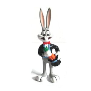 May include: A Bugs Bunny figurine, standing upright, with gray fur and long ears. He wears a black tuxedo, a red bow tie, and holds a carrot and a top hat. The figurine is set against a white background.