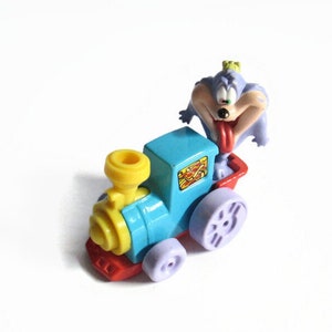 May include: A brightly coloured toy train with a cartoon character on top. The train is mainly blue with yellow and red details. The cartoon character is purple with a long red tongue. The toy has purple wheels.