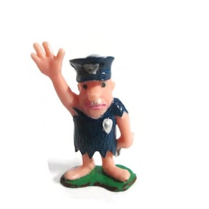 May include: A small plastic figurine of a police officer with peach-colored skin, wearing a dark blue uniform with a jagged hem and a matching cap. The figure has one arm raised in a wave, a silver-colored badge on the chest, and stands on a green base.