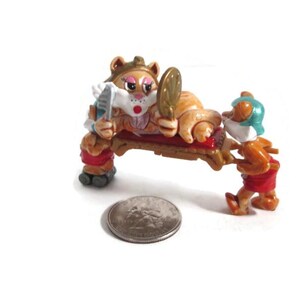 May include: A whimsical figurine depicts a cartoon cat reclining on a wooden bench, holding a mirror and comb. A small figure attends to the cat. The figurine is painted in shades of orange, brown, red, and white. A quarter is next to the figurine.