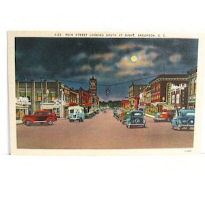 May include: Vintage postcard featuring a nighttime view of Main Street in Anderson, South Carolina. The image shows a street lined with buildings and vintage cars under a moonlit sky. Text on the card reads "A-23 MAIN STREET LOOKING SOUTH AT NIGHT. ANDERSON, S.C."