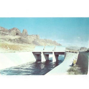 May include: Vintage postcard featuring a concrete bridge over a waterway, with mountains in the background under a blue sky. The water flows beneath the bridge, and a road runs alongside it.