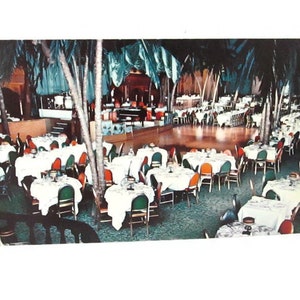 May include: A vintage postcard depicting a large dining room with numerous round tables covered in white tablecloths, surrounded by green chairs. Tall palm trees are integrated into the interior space, and a stage with a piano is visible.