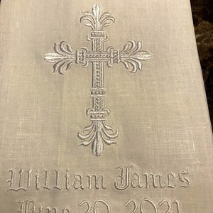 May include: A cream-colored linen cloth featuring an embroidered cross design. The cross is detailed with ornate flourishes. Below the cross, the name "William James" and the date "June 20, 2021" are embroidered in a decorative font.