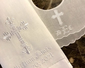 May include: A white baby bib and towel set, both embroidered with religious motifs. The bib features a scalloped edge and a cross. The towel is embroidered with a cross, the name "William James", and the date "June 20, 2021".