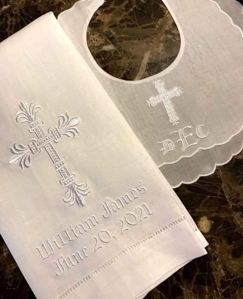 May include: White embroidered baby bib and matching towel set. The bib has a scalloped edge and a cross design. The towel is embroidered with a cross, the name "William James", and the date "June 20, 2021".