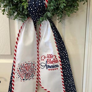 May include: A patriotic wreath with a green leafy base, a navy blue star-patterned ribbon, and white fabric streamers. The streamers are trimmed with red stripes and embroidered with a fireworks design and the words "God Bless America."