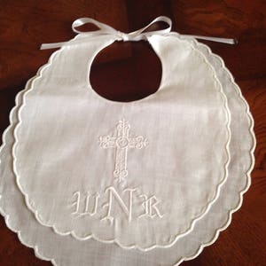 May include: White baby bib with a scalloped edge and a decorative embroidered cross. The bib has a ribbon tie and the initials WNR embroidered below the cross. The bib is made of a soft, white fabric.