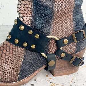May include: A pair of brown and black ankle boots with a snakeskin pattern. The boots have a black leather strap with gold studs and a buckle closure.