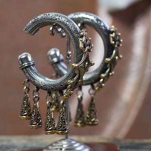 Può includere: Orecchini a cerchio argentati con dettagli dorati e ciondoli a forma di cono pendenti. Esposti su un supporto argentato con base in pietra marrone-rossastra. Il design presenta motivi e texture dettagliate, con uno stile vintage.
