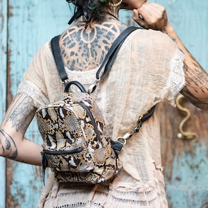 Puede incluir: Una mochila pequeña de piel de serpiente marrón y negra con correas negras. La mochila la lleva una persona con tatuajes.
