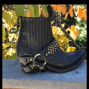 Könnte beinhalten: Schwarze Leder-Stiefeletten mit spitzer Zehenkappe und einem Nietenriemen. Die Stiefeletten haben einen Schnallenverschluss und einen Blockabsatz.