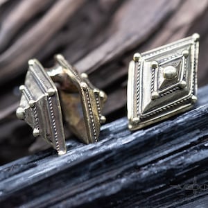 Peut inclure: Gros plan sur deux boucles d'oreilles antiques en laiton avec un motif en forme de losange. Les boucles d'oreilles présentent des détails complexes, notamment une sphère centrale, des bordures en forme de corde et de petits points en relief. Les boucles d'oreilles sont exposées sur une surface sombre et texturée.
