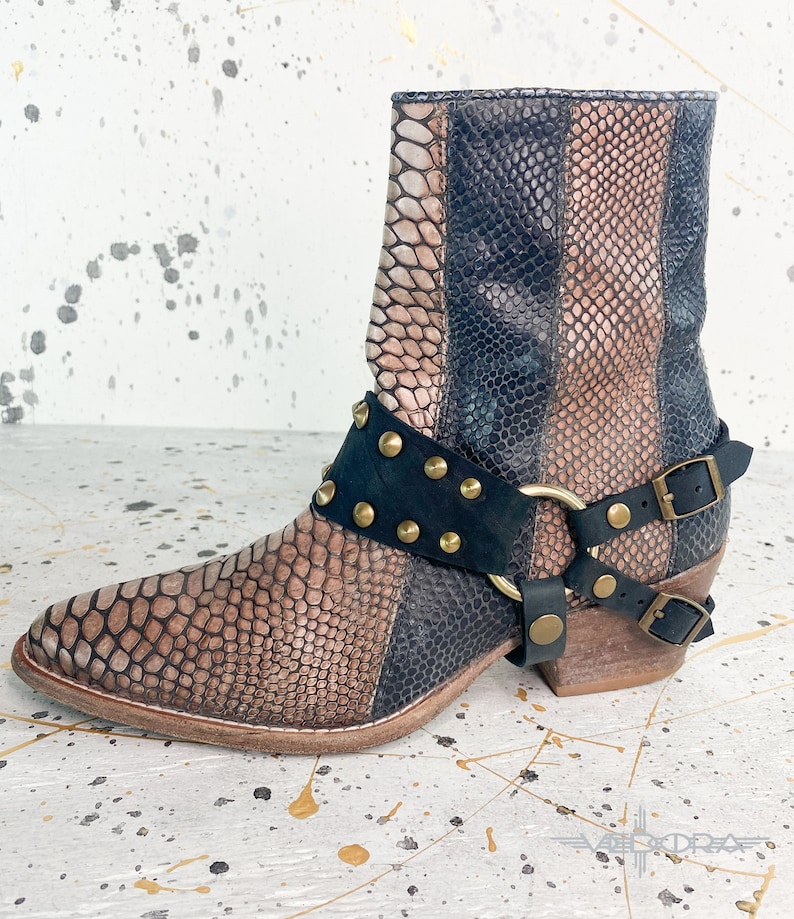 May include: A pair of brown and black ankle boots with a snakeskin pattern. The boots have a black leather strap with gold studs and a buckle closure.