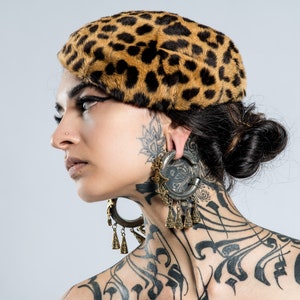 May include: A leopard print beret with a black and brown pattern. The beret is worn with large, ornate earrings and tattoos on the neck and ear. The overall aesthetic is bold and artistic.