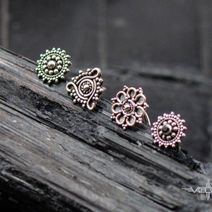 May include: Four silver nose rings with intricate designs. The rings are arranged in a row on a dark wooden surface. The rings are all different shapes and sizes, but they all have a similar ornate style.