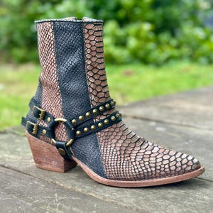 Puede incluir: Botas de tobillo de piel de serpiente negra y marrón con hebillas y tachuelas doradas. Las botas tienen punta puntiaguda y tacón de bloque.