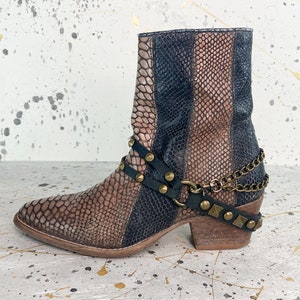 Puede incluir: Botas de tobillo marrones y negras con estampado de piel de serpiente. Las botas tienen un tacón grueso y una correa decorativa con cadena y tachuelas.
