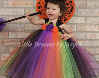 Könnte beinhalten: Ein kleines Kind verkleidet als Hexe für Halloween. Das Kind trägt einen schwarzen und regenbogenfarbenen Tüll-Tutu mit einem schwarzen Oberteil und einen grünen Hexenhut. Das Kind hält einen schwarzen Besen und lächelt.