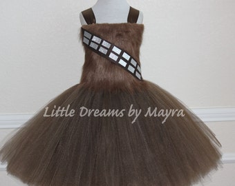 Può includere: Un abito tutu in tulle marrone con un corpetto in pelliccia sintetica e un nastro argentato e nero con un motivo a quadrati ripetuto. L'abito è su un manichino bianco. "Little Dreams by Mayra" è stampato sulla gonna.