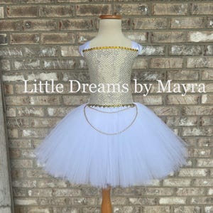 May include: A white tutu dress displayed on a wooden dress form. The bodice is adorned with gold sequins and trim, and a gold chain belt. The text "Little Dreams by Mayra" is printed on the background.