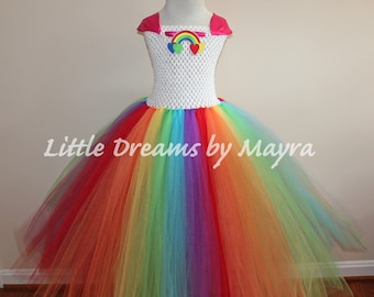 Puede incluir: Un vestido de tutú arcoíris para niñas con un corpiño blanco y un lazo de satén rosa. El corpiño tiene una aplicación de arcoíris y corazones. El tutú está hecho de capas de tul en colores del arcoíris.