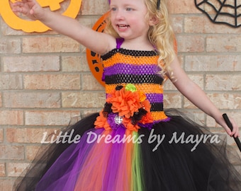 Könnte beinhalten: Ein kleines Mädchen, das sich für Halloween als Hexe verkleidet hat. Sie trägt einen schwarzen Tüll-Tutu mit violetten, orangen und grünen Tüllschichten. Sie hat ein schwarz-orangenes gestreiftes Oberteil mit einer Blumenverzierung. Sie trägt einen lilafarbenen Hexenhut und hält einen Zauberstab.