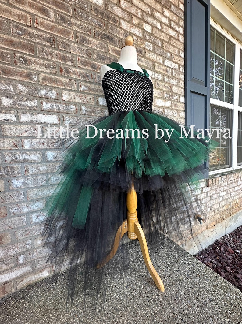 May include: A high-low dress featuring a black crocheted bodice with dark green velvet straps and a voluminous layered tulle skirt in alternating black and dark green colors. The text Little Dreams by Mayra is visible across the center.